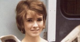 A woman with short brown hair and hoop earrings stands in front of a blue and white door, smiling softly at the camera.