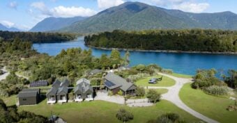 A group of cabins and buildings sit by a bright blue lake, surrounded by green trees and mountains under a partly cloudy sky. A small helicopter is parked on a grassy clearing near the water.