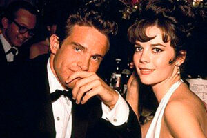 A man in a tuxedo and a woman in a light blue dress sit close together at a formal event, both smiling at the camera. The background shows dim lighting and other guests.