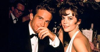 A man in a tuxedo and a woman in a light blue dress sit close together at a formal event, both smiling at the camera. The background shows dim lighting and other guests.