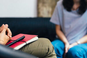 Two people sit facing each other on a couch. One person holds a red notebook and pen, appearing to listen, while the other sits with hands in their lap, suggesting a counseling or therapy session.