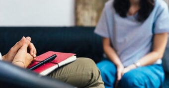 Two people sit facing each other on a couch. One person holds a red notebook and pen, appearing to listen, while the other sits with hands in their lap, suggesting a counseling or therapy session.