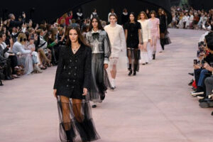 Models walk down a runway wearing stylish outfits, including black, white, and pink dresses, with a seated audience on both sides taking photos during a fashion show.