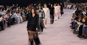 Models walk down a runway wearing stylish outfits, including black, white, and pink dresses, with a seated audience on both sides taking photos during a fashion show.