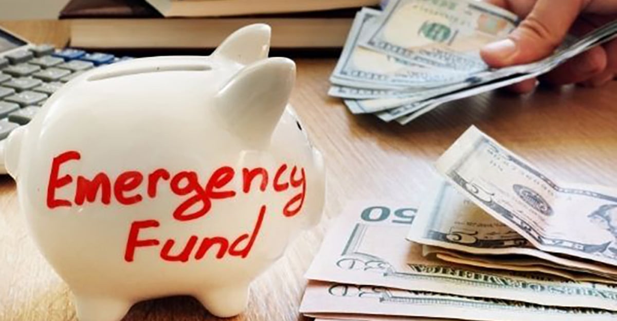 A white piggy bank labeled "Emergency Fund" sits on a table with scattered US dollar bills, while a person counts more cash in the background.