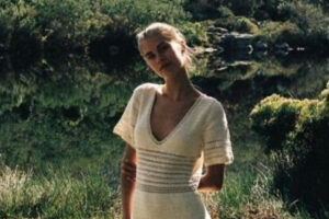 A woman in a white, short-sleeved dress stands outdoors near a pond, surrounded by greenery and trees, with sunlight creating a serene and natural atmosphere.