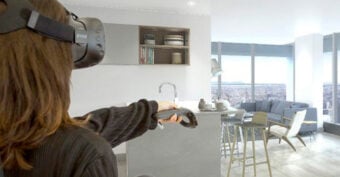 A person wearing a VR headset and holding a controller stands in a modern, bright apartment kitchen with large windows and city views.