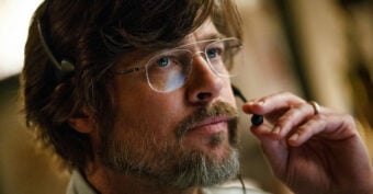 A man with brown hair, a beard, and glasses wears a headset with a microphone, gazing thoughtfully ahead while holding the microphone near his mouth.