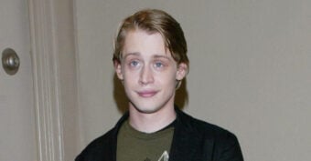 A young man with light skin and blond hair, wearing a dark jacket over a green shirt, stands indoors against a plain wall, looking at the camera with a slight smile.