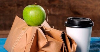 A green apple rests on two brown paper-wrapped packages tied with string, next to a white disposable coffee cup with a black lid on a blue surface and a brown background.