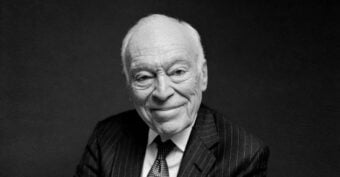 An elderly man in a dark pinstripe suit and tie sits against a plain, dark background, smiling gently at the camera.