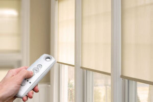 A hand holds a remote control, adjusting beige roller blinds on large windows in a sunlit room.