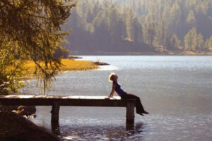 A person sits relaxed at the edge of a wooden dock, overlooking a calm lake surrounded by trees and forested hills, with sunlight filtering through the scene.