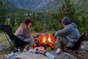 Two people sit in camping chairs by a campfire in a grassy, mountainous area, surrounded by pine trees and camping gear, with a scenic valley and mountains in the background.