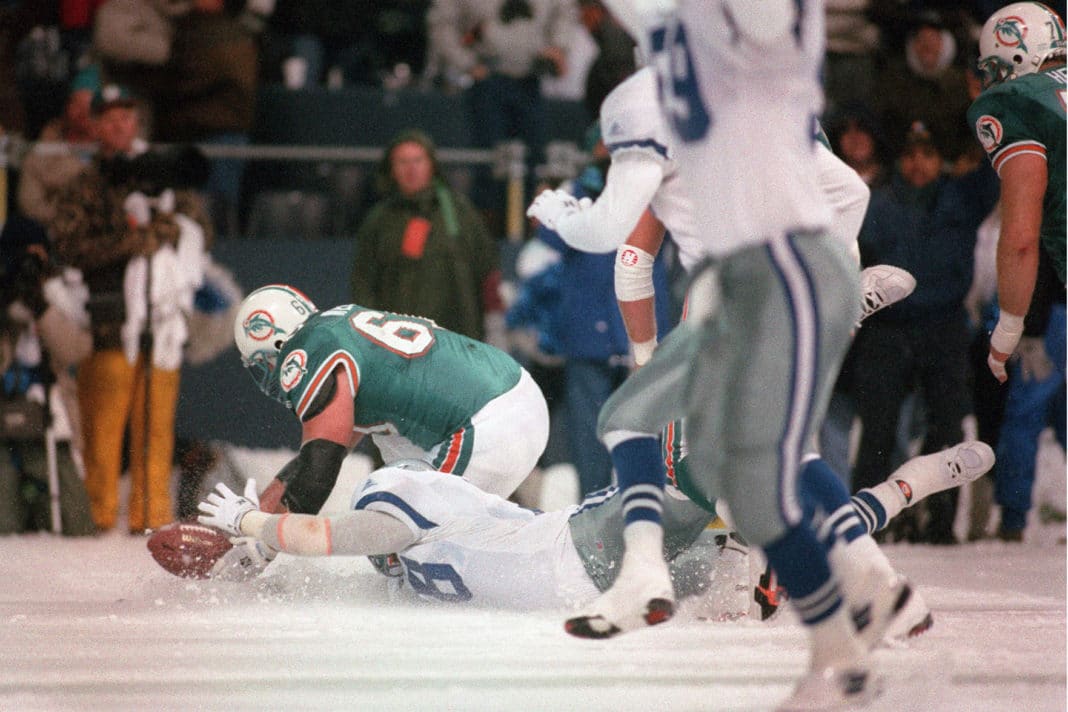 A football player in a white uniform dives for a loose ball on a snow-covered field, while a player in a green Miami Dolphins uniform attempts to recover it. Other players and spectators are visible in the background.