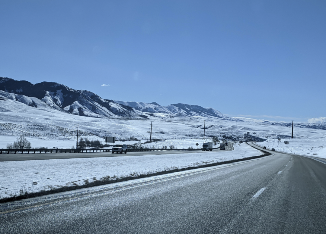 A two-lane highway curves through a snowy landscape with mountains in the background. Several vehicles travel in both directions under a clear blue sky. Snow covers the fields and hills along the roadside.