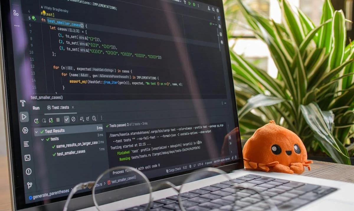 A laptop displaying code and test results in an IDE sits on a desk next to a small orange plush toy and a green leafy plant in the background. Career.