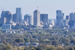 A clear daytime view of a city skyline with modern high-rise buildings and skyscrapers, surrounded by trees and residential neighborhoods in the foreground.