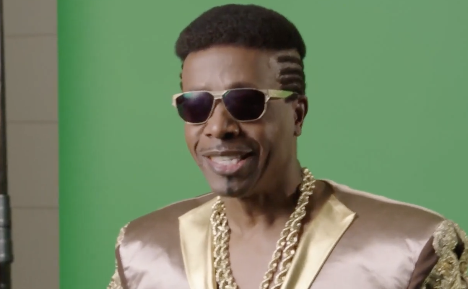 A man wearing sunglasses, gold chains, and a shiny gold jacket stands in front of a green background. He has a styled haircut with shaved designs on the side and is smiling.