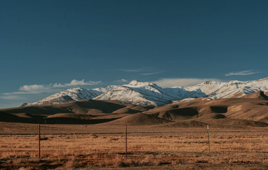 Snow-capped mountains rise under a clear blue sky, with rolling brown hills and dry grassland in the foreground, separated by a simple wire fence.