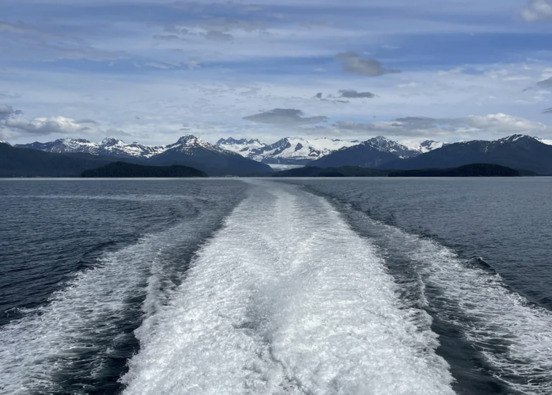 A boat’s wake cuts through calm blue water, leading toward distant snow-capped mountains under a partly cloudy sky. Forested hills line the horizon.