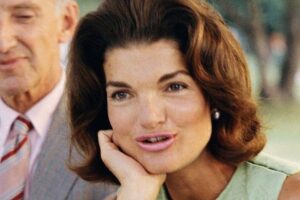 A woman with medium-length brown hair wearing a sleeveless light green top rests her chin on her hand and looks forward. An older man in a suit and striped tie stands beside her, slightly out of focus.
