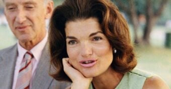 A woman with medium-length brown hair wearing a sleeveless light green top rests her chin on her hand and looks forward. An older man in a suit and striped tie stands beside her, slightly out of focus.