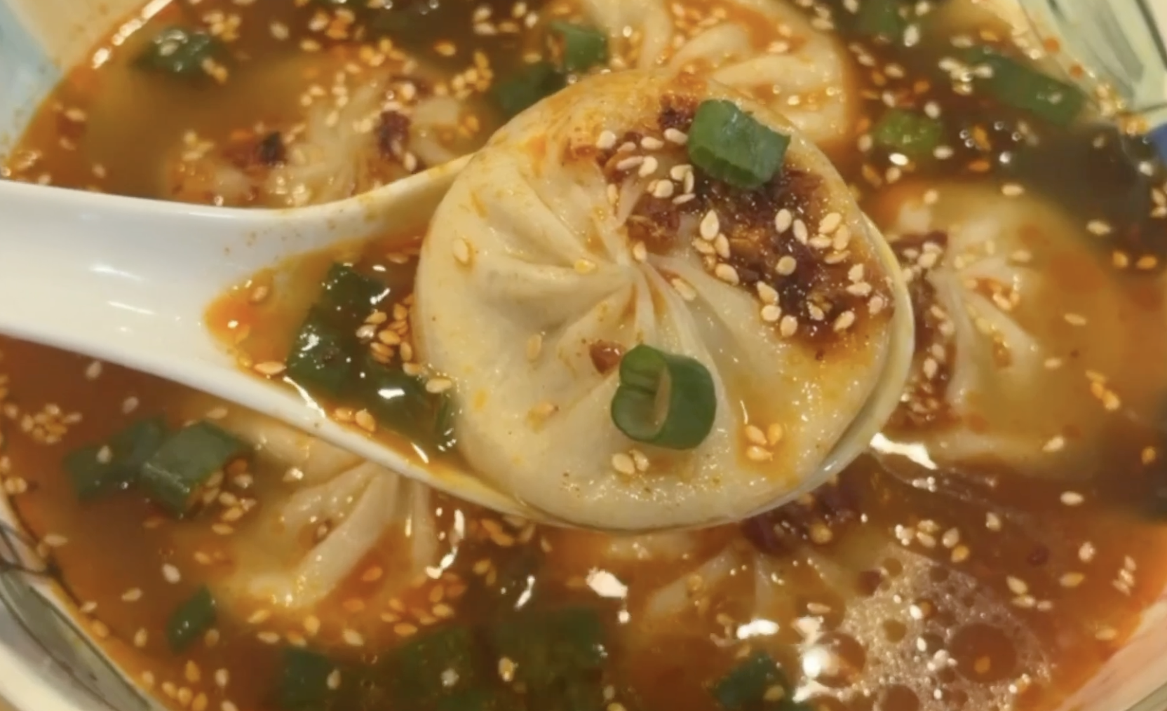 A soup dumpling held in a white spoon over a bowl of spicy red broth topped with sesame seeds and green onions.