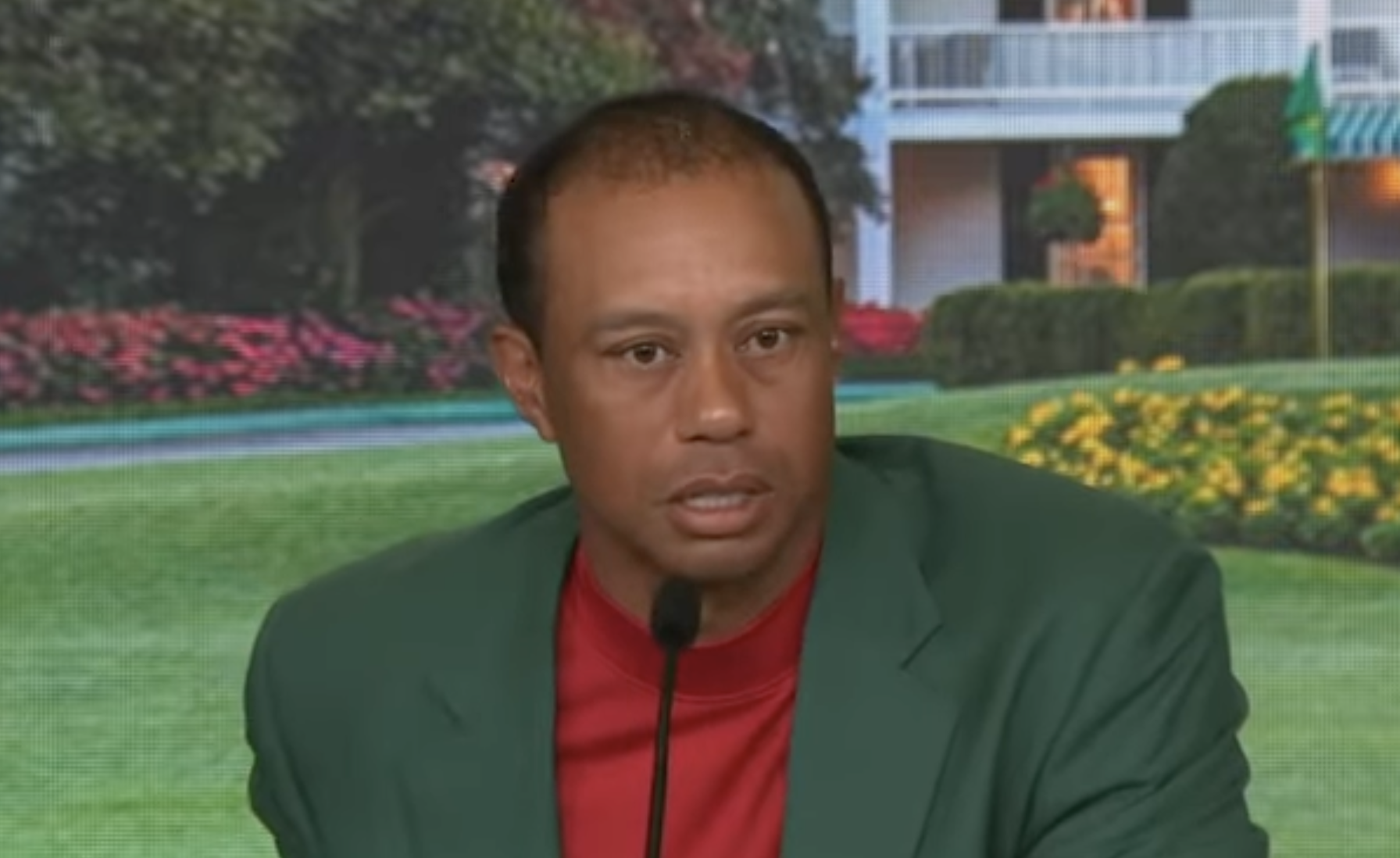 A man wearing a green jacket and red shirt speaks into a microphone at a press conference, with a backdrop of a building, green lawn, and flowerbeds behind him.