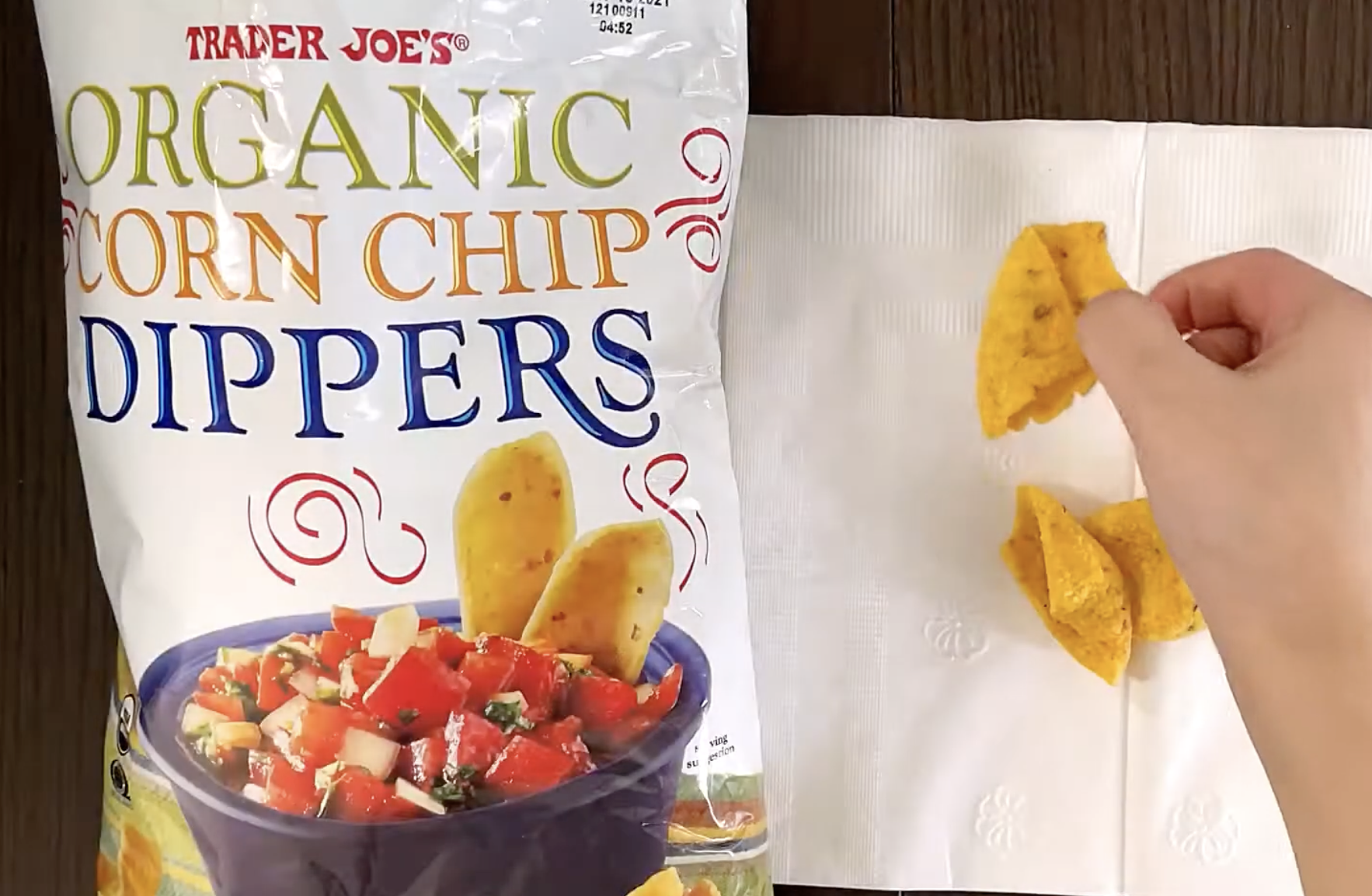 A hand places Trader Joe’s Organic Corn Chip Dippers onto a white paper napkin next to the product bag.