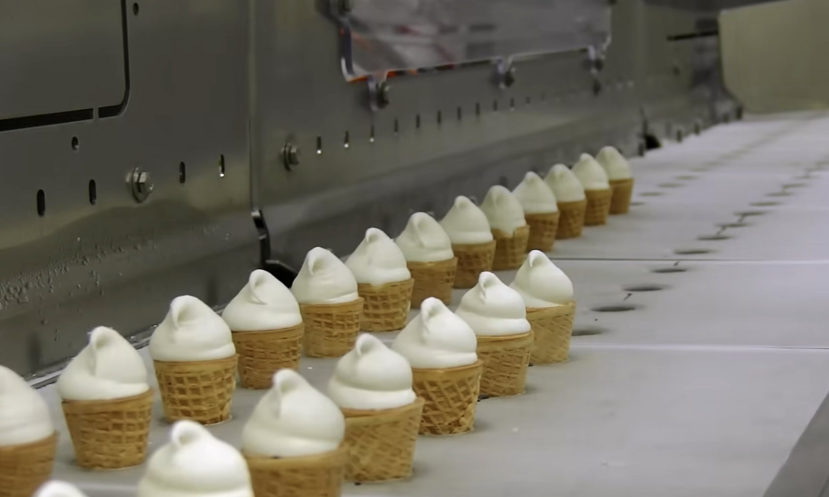 Rows of mini ice cream cones with soft-serve swirls move along a factory conveyor belt.