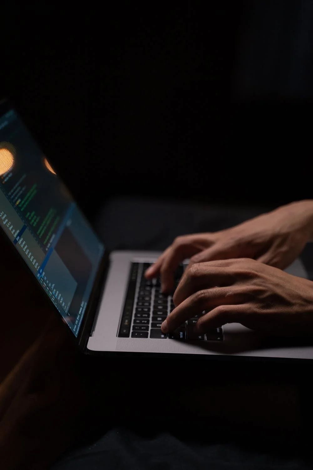 Hands typing on a laptop keyboard in a dimly lit setting, with code visible on the laptop screen.