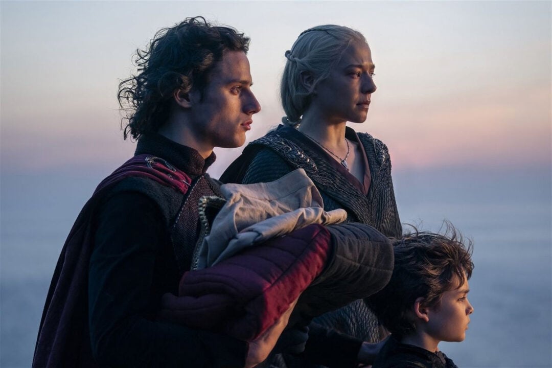 Three people stand together at sunset, looking into the distance. One holds a bundle of blankets, another stands close by, and a child is in front of them. All appear serious and contemplative.