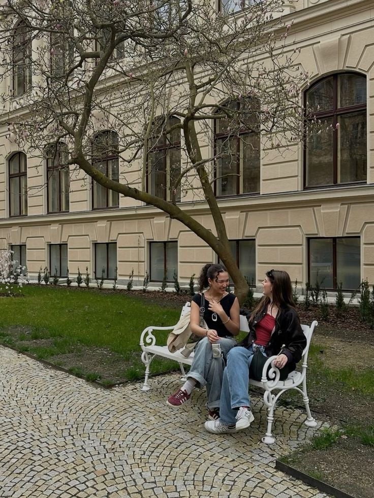 Two women sit on a white bench under a budding tree, chatting and smiling. They are in front of a cream-colored building with large windows and surrounded by cobblestone and grass.