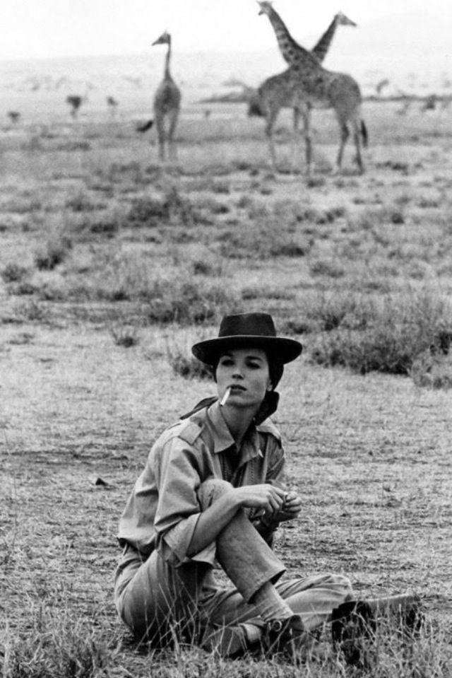 A woman in safari clothing and a hat sits on dry grass, holding a cigarette, with giraffes walking in the background on an open plain. The scene appears calm and natural.