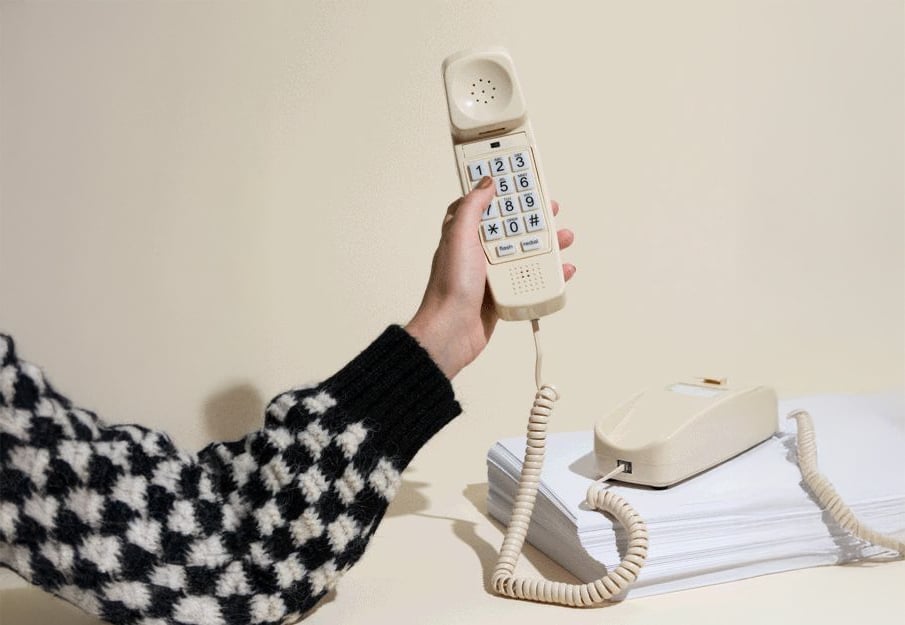 A person wearing a black-and-white checkered sweater holds the handset of a beige corded landline phone, which rests on a stack of white papers against a plain light background.