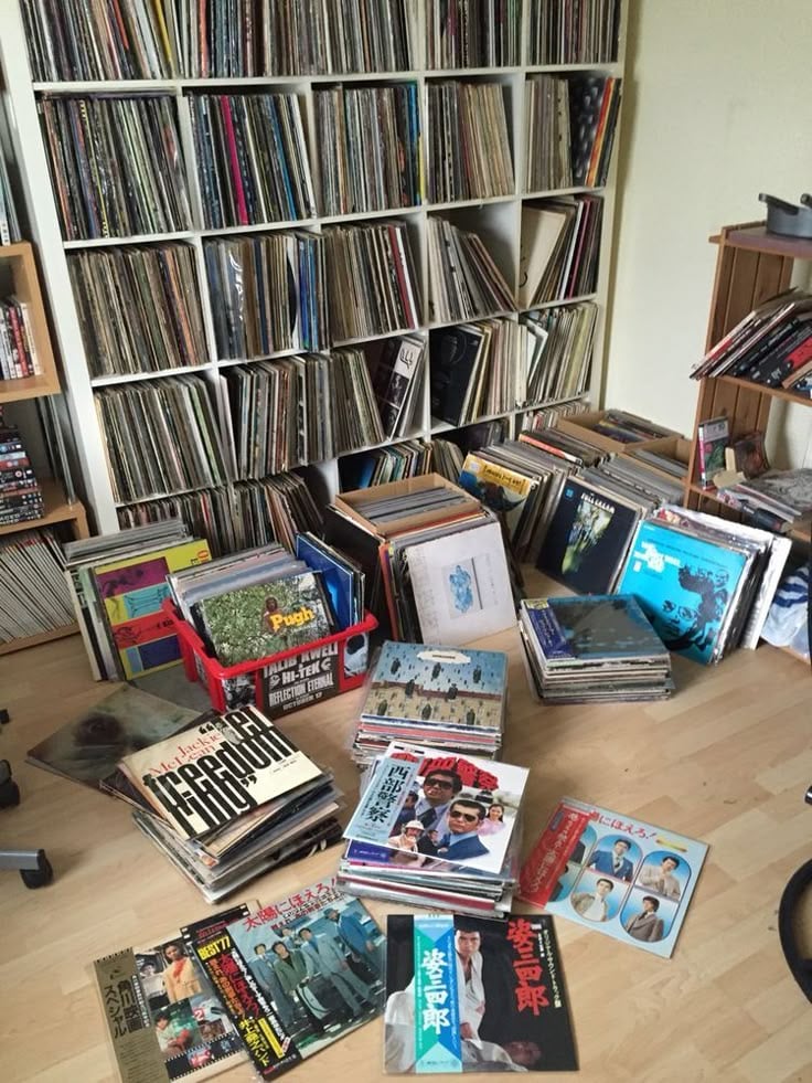 A room with shelves full of vinyl records and many more records stacked and spread on the wooden floor, featuring colorful album covers in various languages and styles.