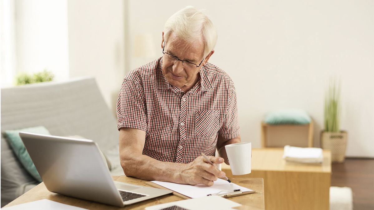 An older man with gray hair and glasses sits at a table, holding a white mug and writing on a notepad while looking at a laptop screen in a bright, modern living room.