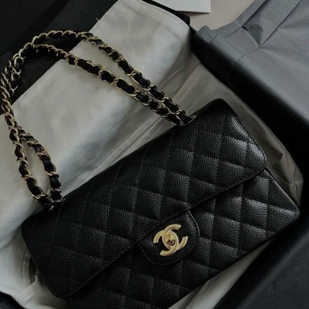 A black quilted leather Chanel handbag with a gold chain strap and gold interlocking CC logo clasp, resting on white fabric inside a box.