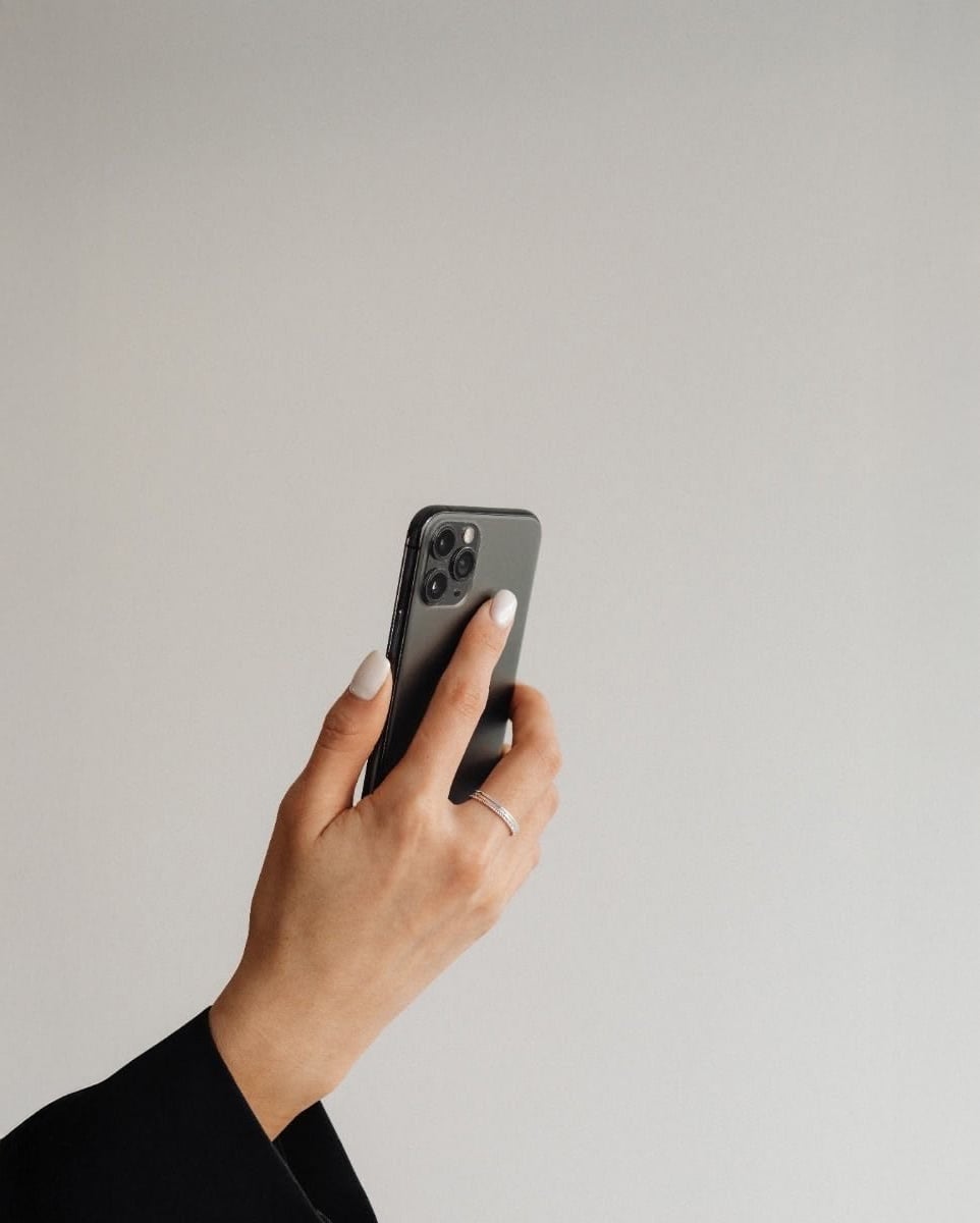 A person with light skin and manicured nails holds a black smartphone with three cameras, wearing a black sleeve and a silver ring, against a plain light gray background.