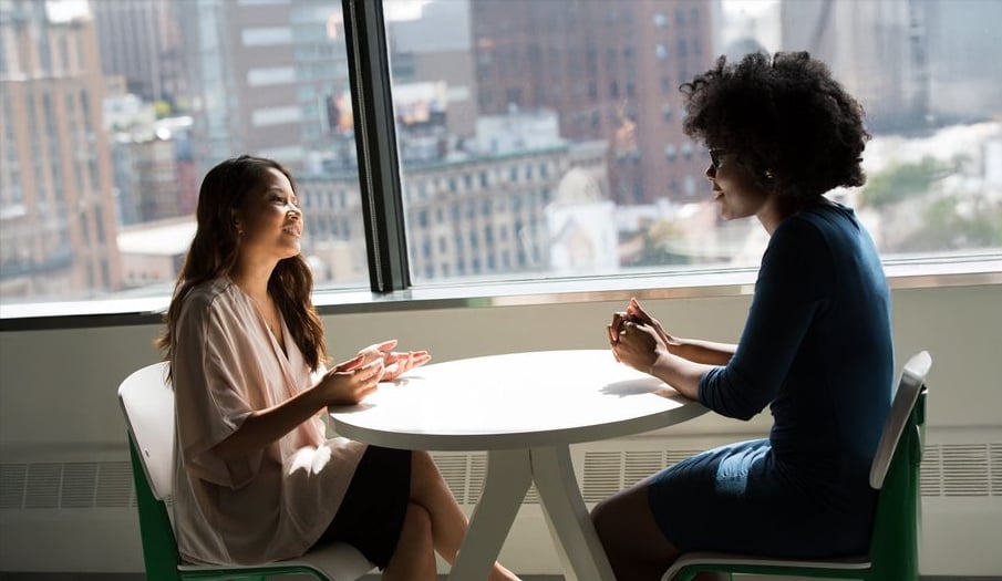 Two women sit across from each other at a round white table by a large window, engaged in conversation. City buildings are visible outside, and natural light fills the room.
