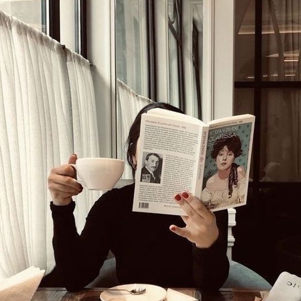 A person in a black top sits by a window, holding a large white cup in one hand and reading a book titled "Clarissa" by Stefan Zweig, with their face mostly hidden behind the book.