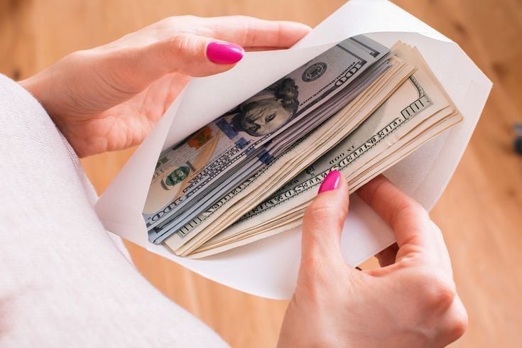 A person with pink nail polish holds an open white envelope filled with a large stack of U.S. hundred-dollar bills.