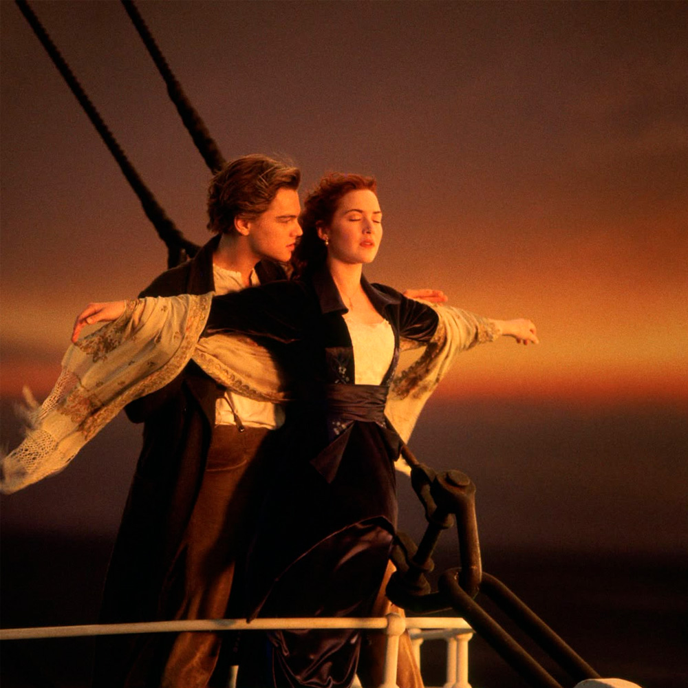 A man and woman stand on the bow of a ship at sunset, with the woman spreading her arms wide and the man holding her from behind. They appear joyful and windblown, with the ocean and orange sky in the background.