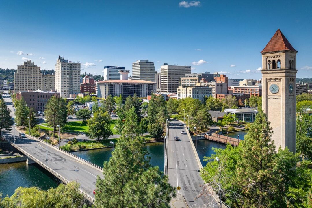 A scenic view of a city skyline with tall buildings, a clock tower, a bridge over a river, and lush green trees under a clear blue sky.