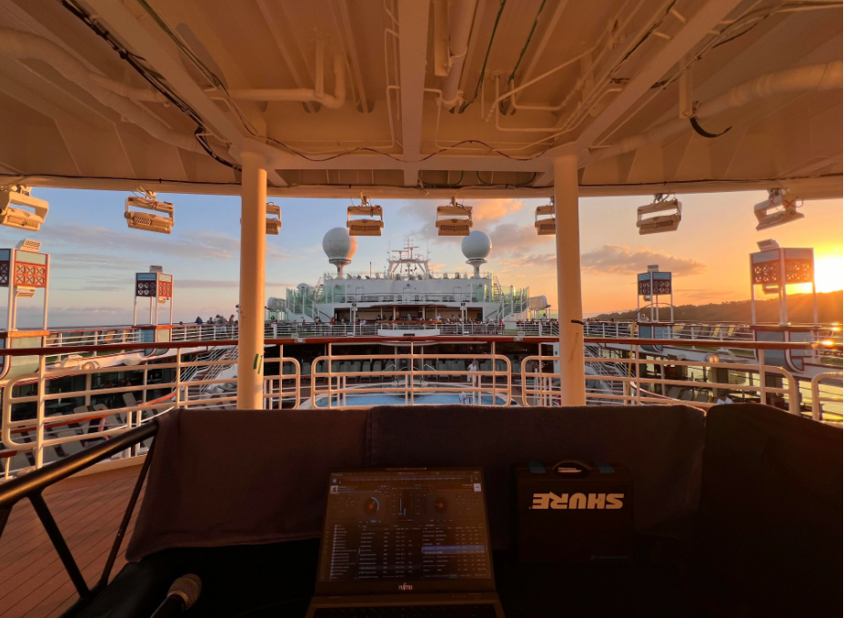 A view from a shaded upper deck on a cruise ship at sunset, overlooking the pool area and the upper decks, with lighting equipment overhead and a DJ booth with a mixer in the foreground.