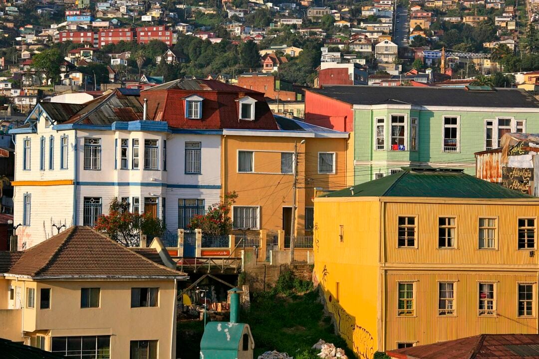 Colorful houses with varied architecture are built on a hillside in a densely populated area, with greenery and more buildings in the background under warm sunlight.