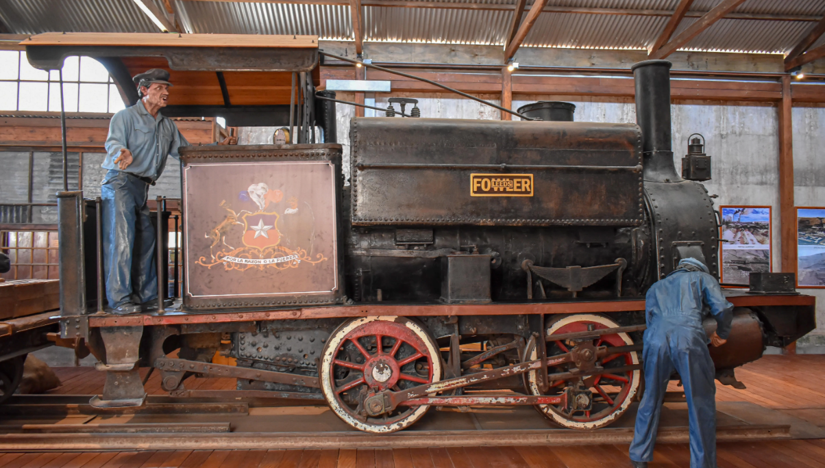A vintage black steam locomotive is displayed indoors, with two mannequins dressed as railway workers positioned on and beside the train. The setting features wooden floors and a corrugated metal roof.