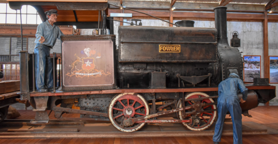 A vintage black steam locomotive on display indoors, with two realistic human mannequins posed as workers&mdash;one standing on the train, the other inspecting a wheel. The locomotive sits on wooden flooring.