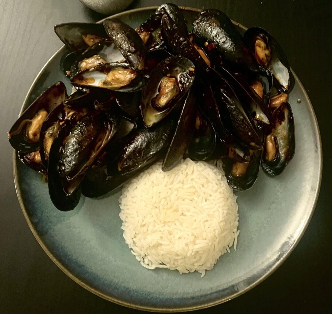 A plate with cooked mussels in dark shells and a neat mound of white rice on a dark surface, with a grey stone placed above the plate.